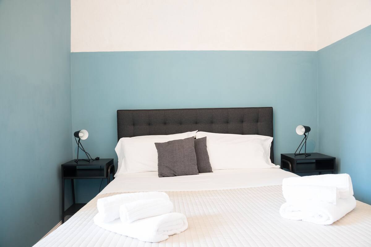 Una cama bien hecha con sábanas blancas y dos almohadas, dos mesitas de noche con lámparas, y una pared azul.