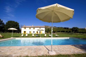 Une piscine entourée de chaises longues et d’un parasol, elle-même bordée d’une terrasse en pierre et, en arrière-plan, d’un grand bâtiment blanc agrémenté de touches de jaune.