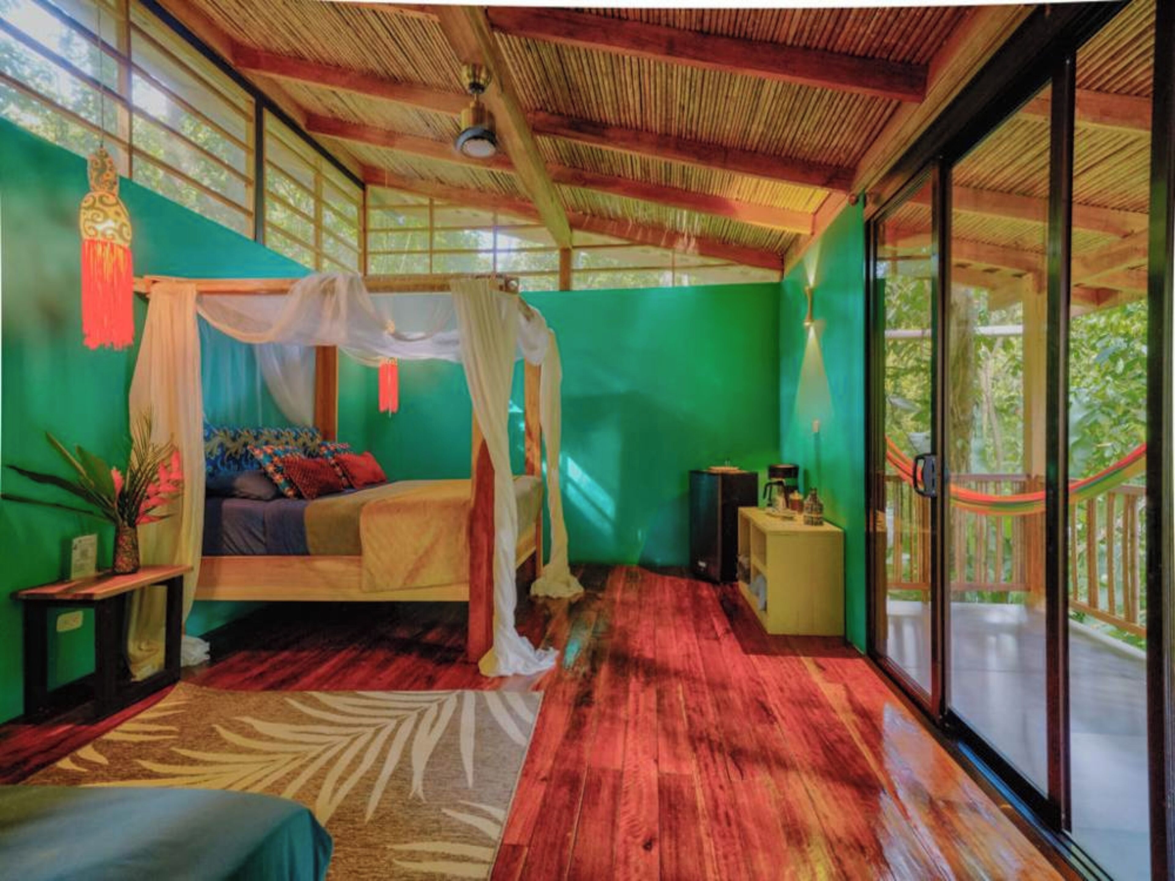 A room with a bed under a canopy, a small table with a lamp, and a view of the outdoors through large windows.