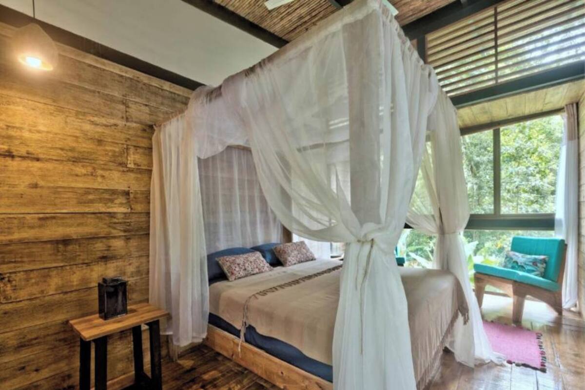 Una habitación de madera con una cama bajo un dosel, un banco y una ventana con vista a follaje.