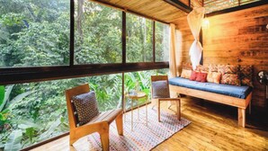 Interior de madera con un amplio ventanal que da a un bosque, un banco con cojines de colores y dos sillas.