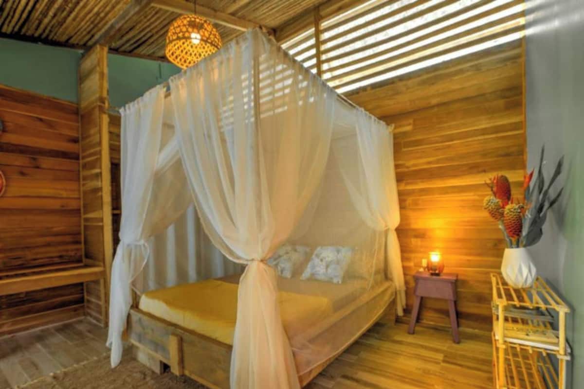 Una habitación de madera con una cama bajo un dosel, una mesita de noche pequeña con una lámpara y una repisa de pared con adornos.