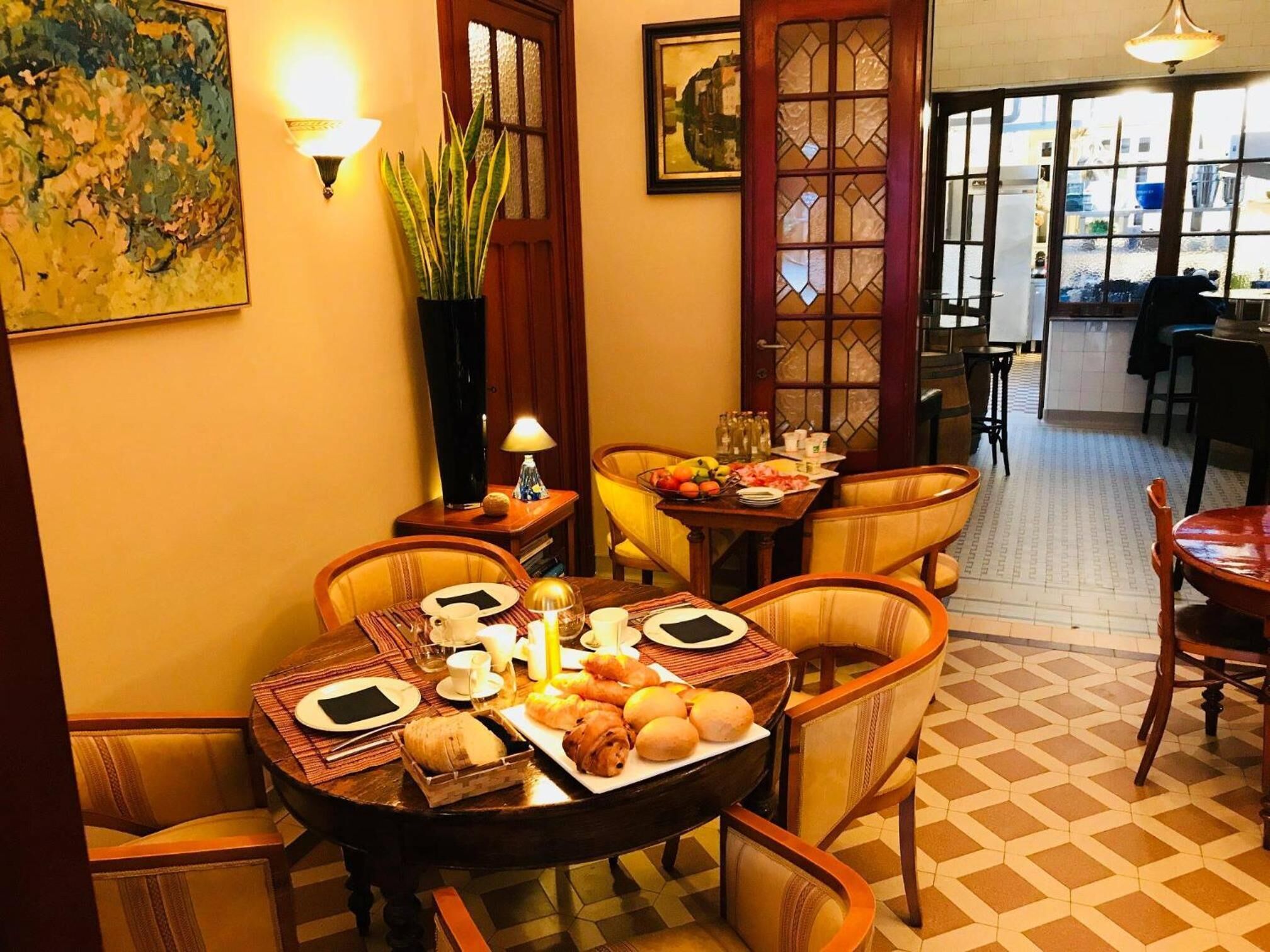 Un coin repas avec une table ronde dressée pour quatre personnes, entourée de chaises rembourrées. On y trouve des assiettes, des tasses et une variété de pâtisseries.