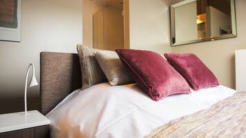 A neatly made bed with white linens and three pillows, a bedside lamp, and a mirror on the wall.