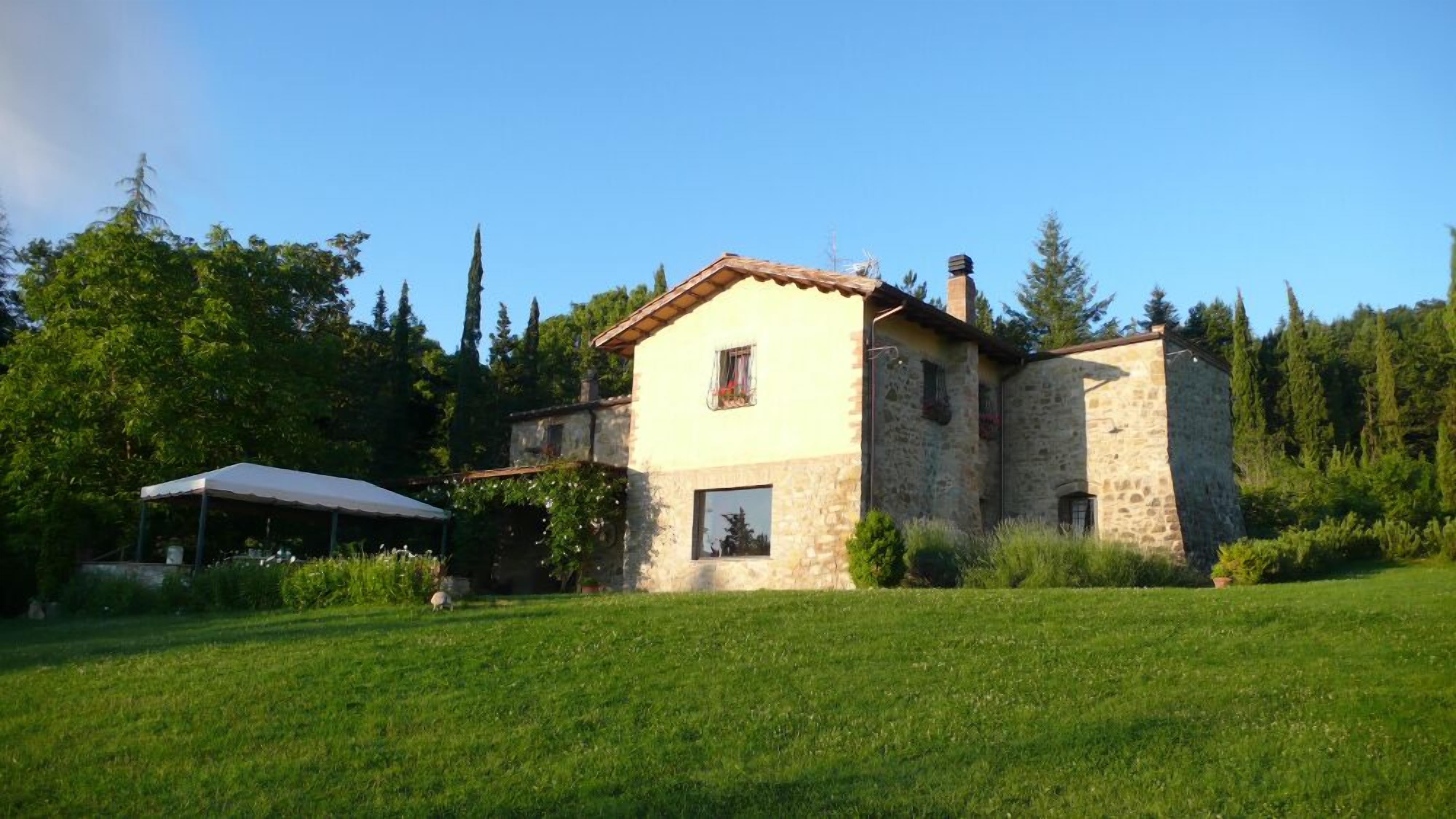 Une maison en pierre avec un toit de tuiles, entourée d’un champ herbeux et d’arbres.
