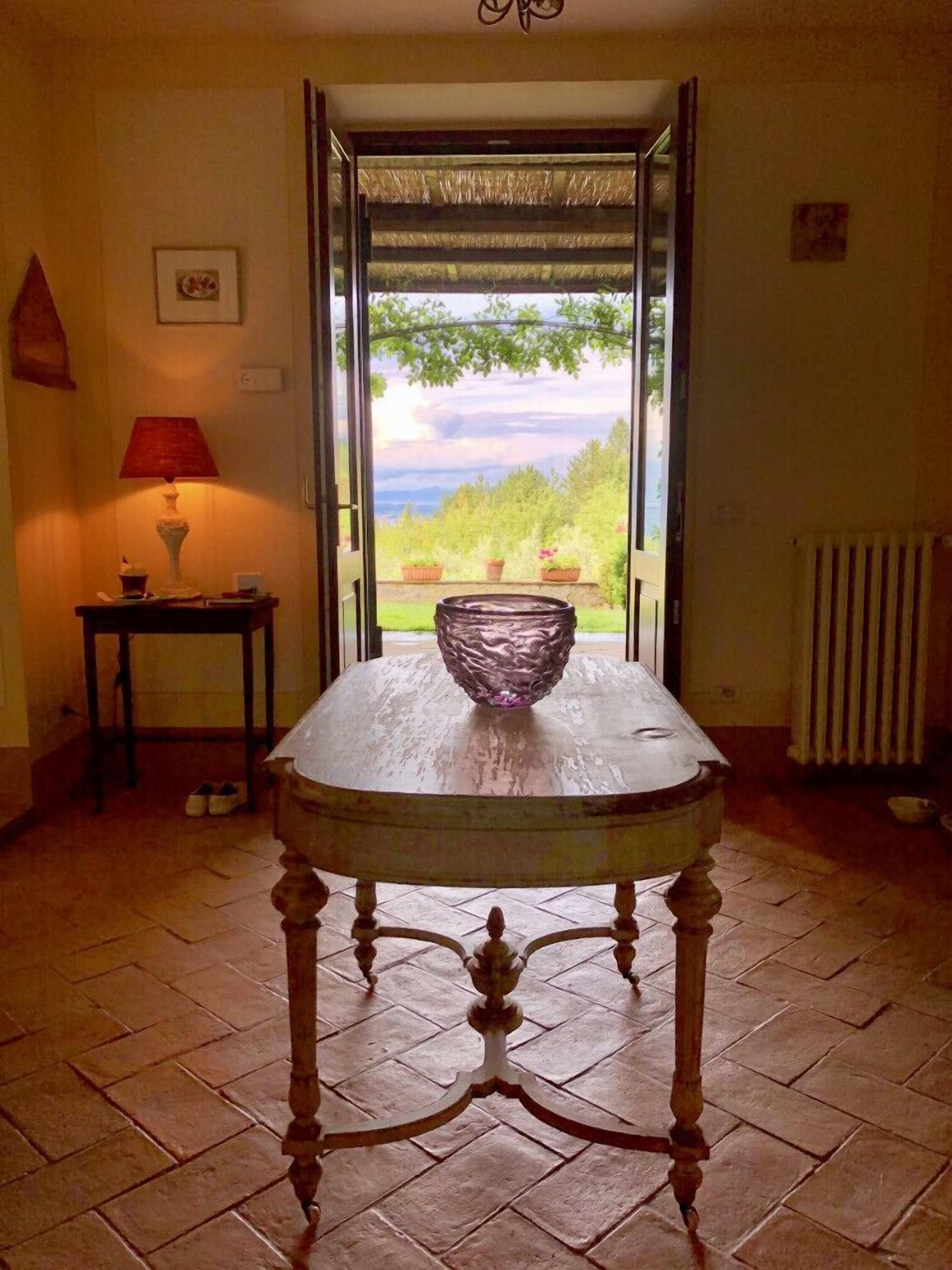 A table with a bowl in the center, a lamp on a side table, and a view of greenery through an open door.