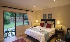 A bedroom with a bed, two bedside lamps, a nightstand, a desk, a chair, and a view of the garden through the window. - SomerZicht Guesthouse (Cape Town)