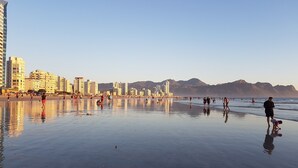 Een breed strand met mensen die wandelen en spelen, gebouwen op de achtergrond en bergen in de verte.