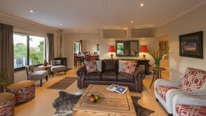 Een ruime woonkamer met een bruine leren bank, een witte bank met gestreepte kussens, een houten salontafel en een eethoek op de achtergrond.