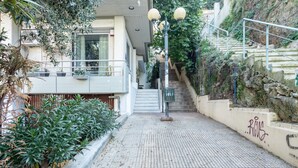 Un chemin pavé mène à un escalier doté d’une rampe, le tout étant entouré de verdure et bordé par un bâtiment avec un balcon.