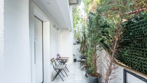 Un étroit couloir extérieur agrémenté de plantes en pot, un espace détente avec des chaises et des tables, et un mur blanc d’un côté.