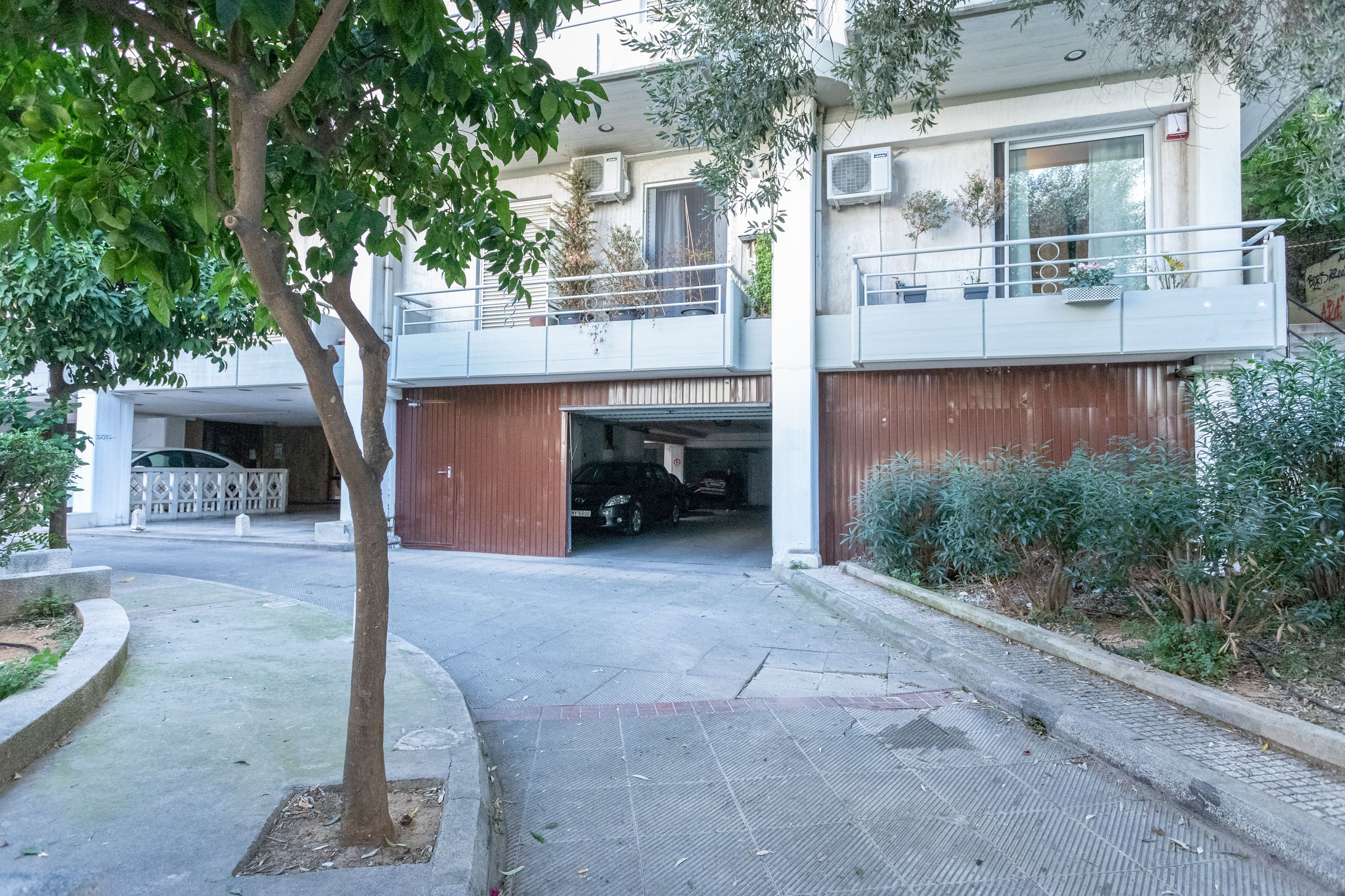 A modern two-story residential building with a balcony, a garage, and a tree in the foreground.