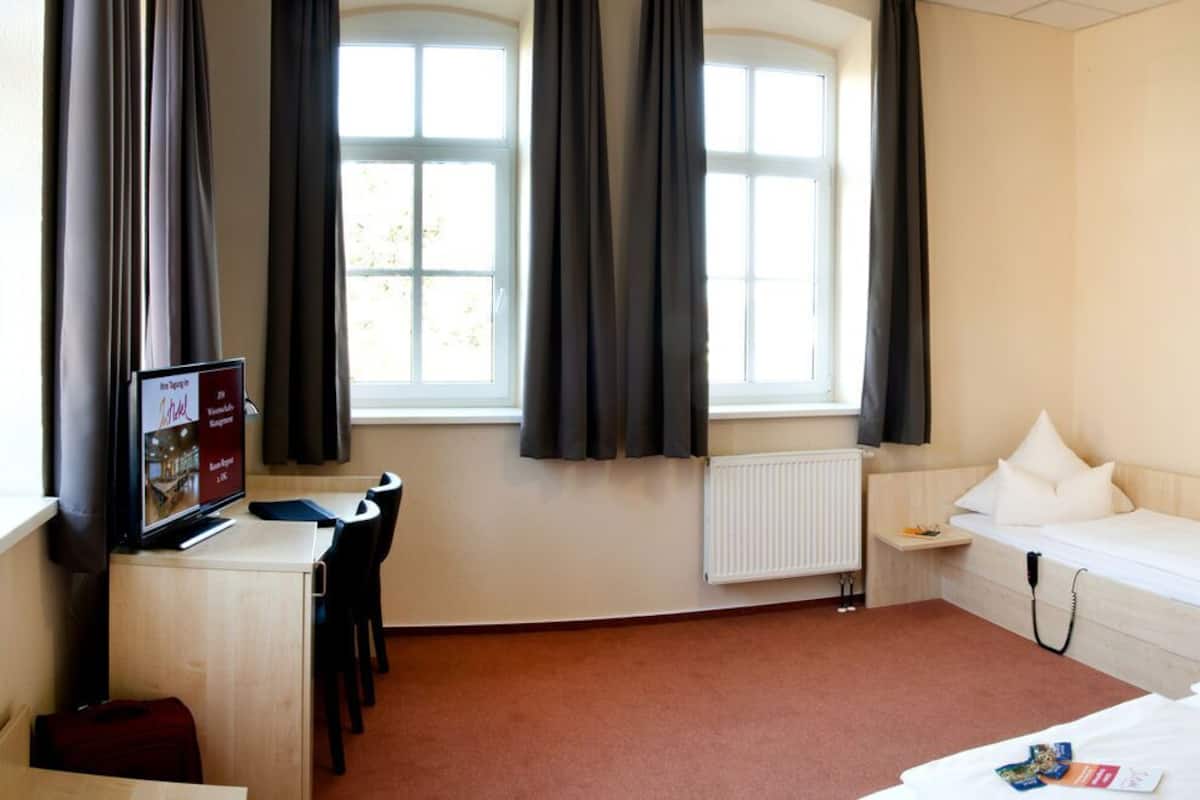 A hotel room with a bed, a desk with a computer, a chair, a window with curtains, a radiator, and a door with a mirror and notices.