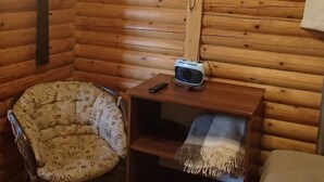 Une pièce chaleureuse aménagée dans un chalet en bois, avec un téléviseur fixé au mur, une petite table en bois sur laquelle est posée une enceinte, un fauteuil à motifs et un banc avec une couverture.