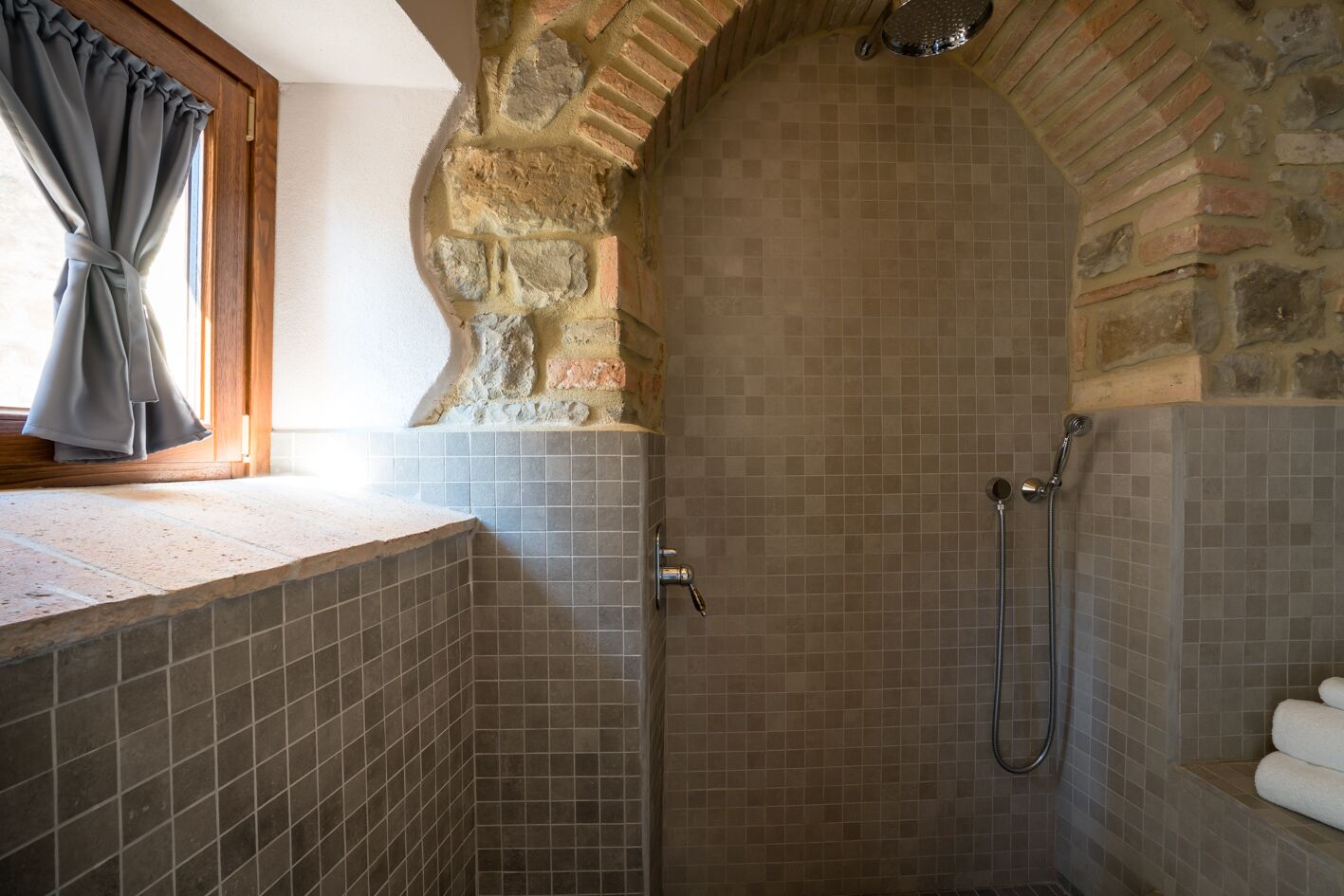 Une douche avec des murs en pierre et un sol carrelé, une fenêtre avec des rideaux et un porte-serviettes.