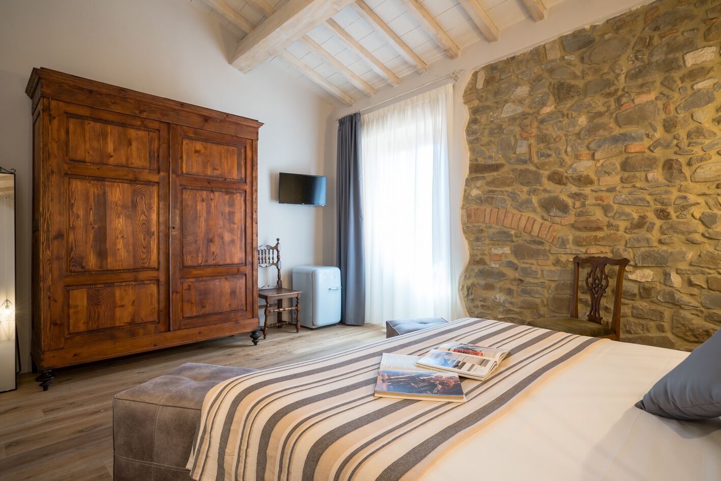 A bedroom with a large wooden wardrobe, a stone wall, a bed with striped bedding, and a small table with a book.