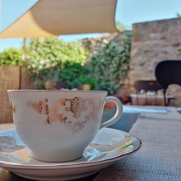 Una taza de cerámica blanca con detalles dorados sobre un platito, colocada sobre una mesa al aire libre, con un muro de piedra y follaje en el fondo.