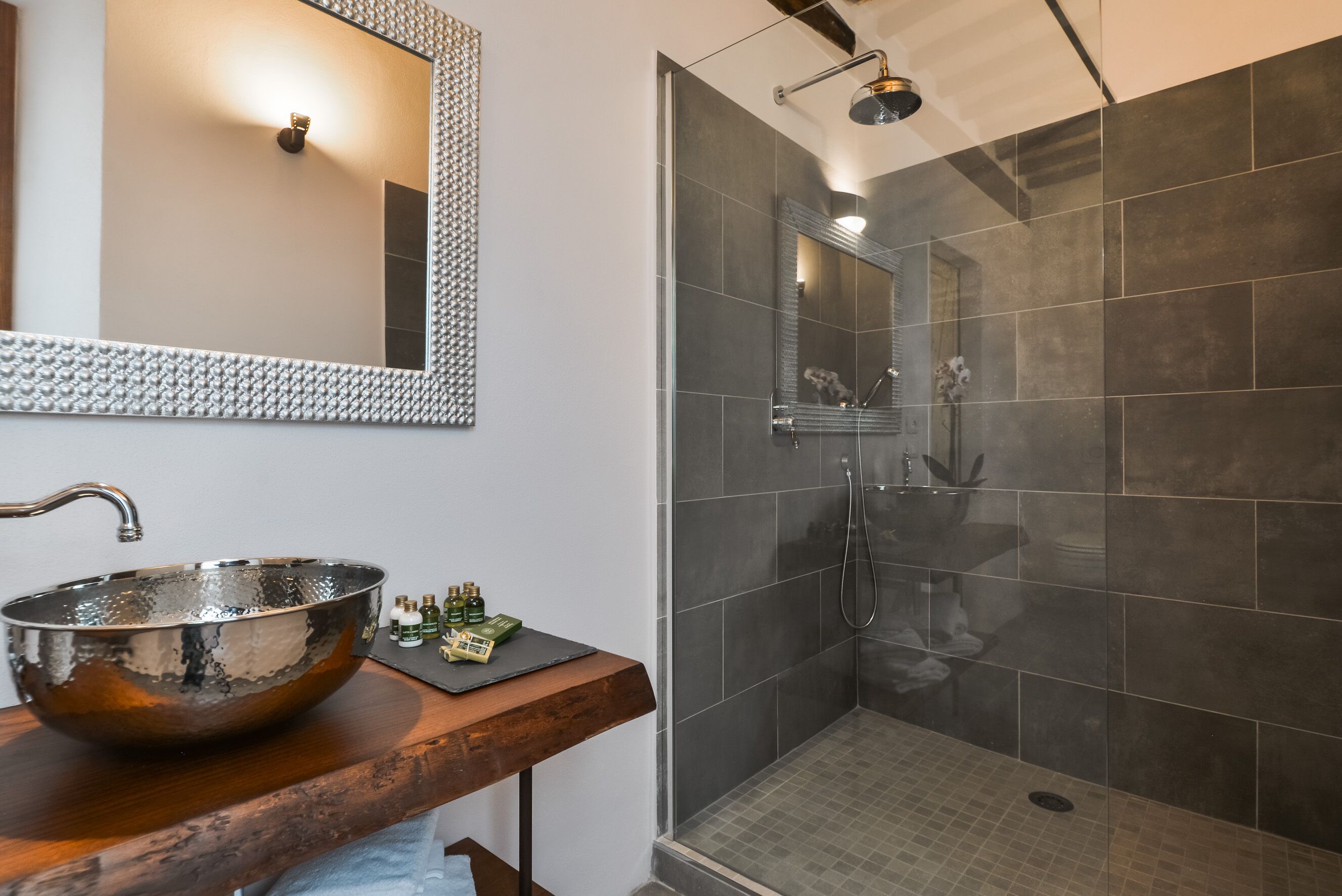 A bathroom with a large, reflective vessel sink, a wooden countertop, a mirror with a textured frame, and a glass-enclosed shower area with dark tiles.