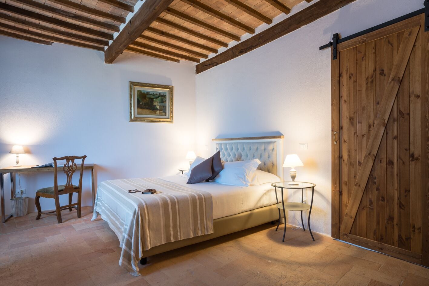 Un dormitorio con cama, mesitas de noche, una silla, un escritorio y una puerta de madera.