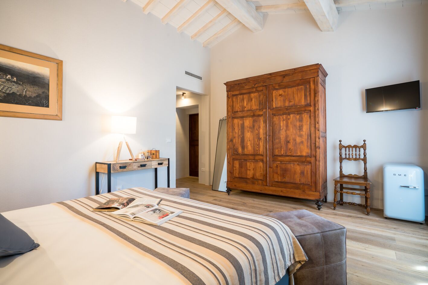 A bedroom with a bed, a wooden wardrobe, a desk with a lamp, a chair, and a small refrigerator.