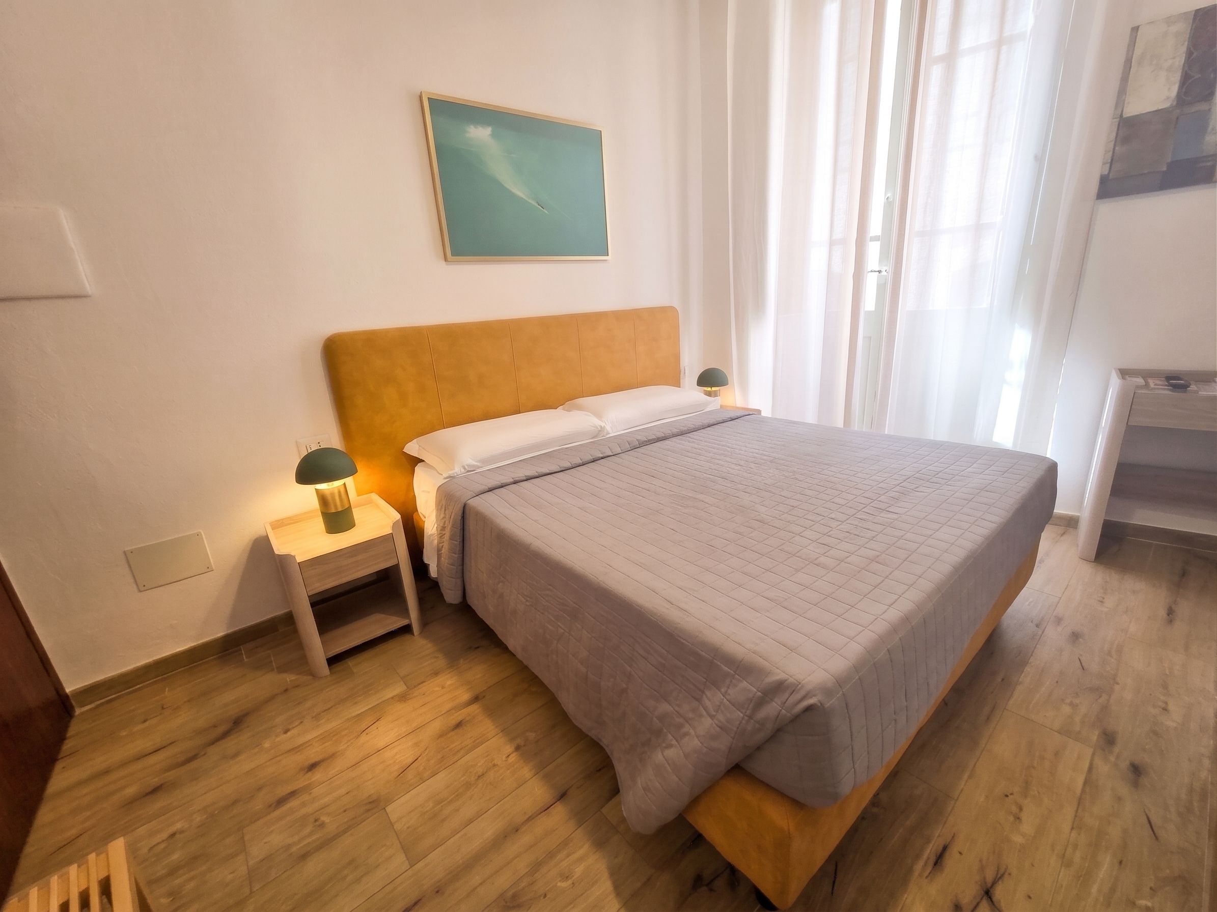 A bedroom with a wooden bed, a nightstand with a lamp, a painting on the wall, and a window with sheer curtains.