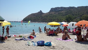 A beach with people sunbathing and swimming, umbrellas, and a mountain in the background. - Le Tre Stelle (Cagliari)