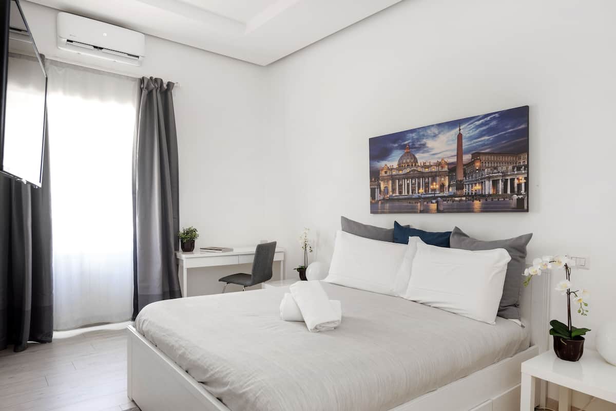 A modern hotel room with a large bed, a desk, and a chair. There is a painting of a cityscape on the wall.