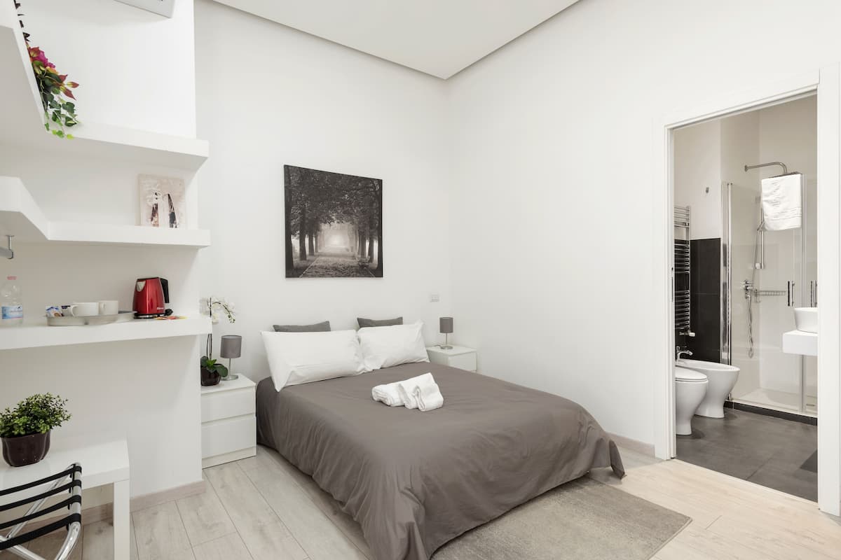 A modern bedroom with a bed, bedside tables, a nightstand, a coffee maker, a potted plant, a framed picture, and a bathroom with a shower and toilet.