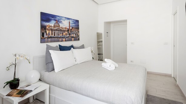 A modern bedroom with a large bed, white bedding, a nightstand with a lamp, a framed picture of a building, and a potted plant.