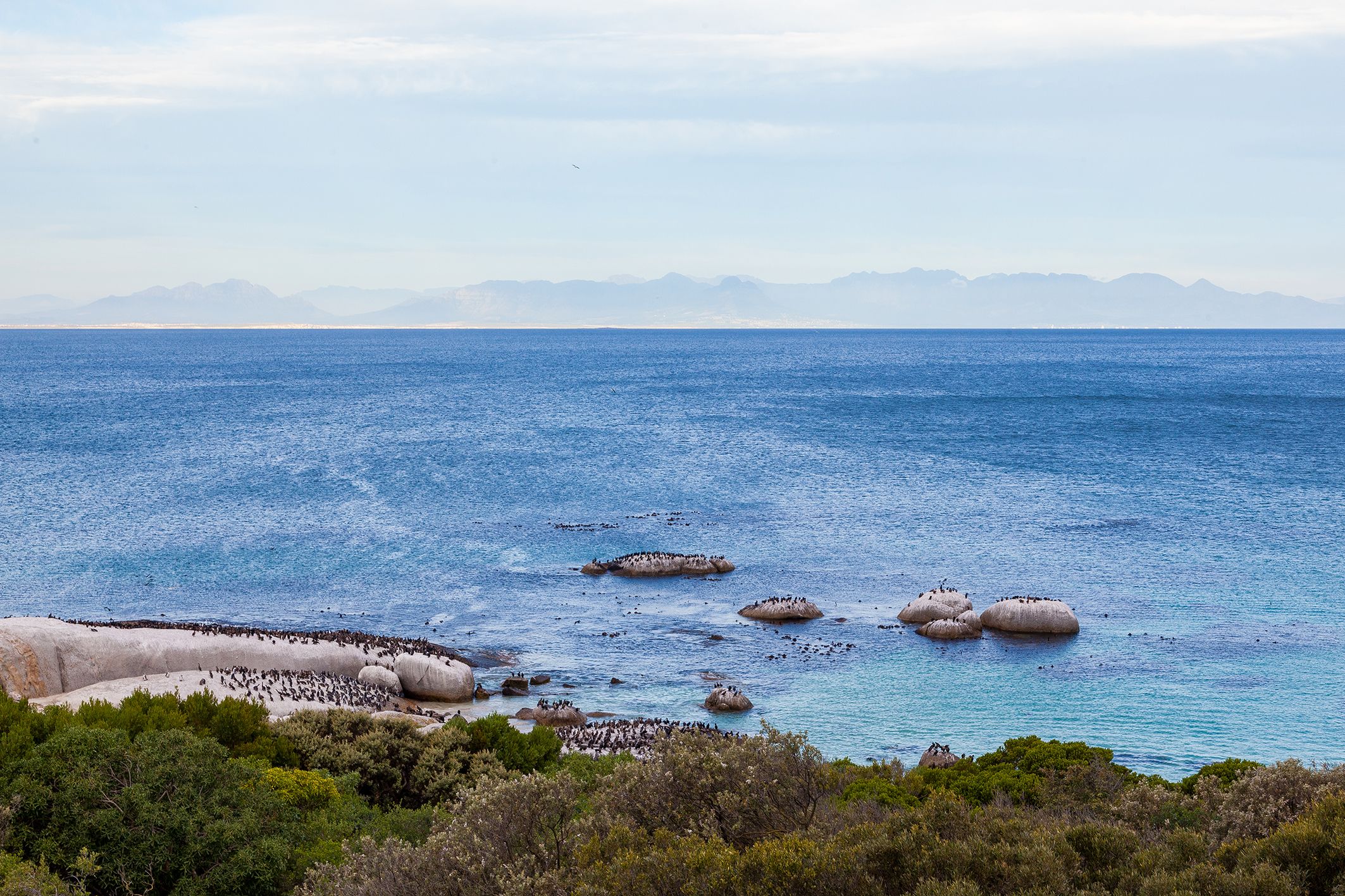 A coastal landscape with clear blue sea, rocky outcrops, and distant mountains.