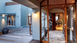 Un espacio al aire libre cubierto con un banco de madera, una silla de mimbre y un ventanal de vidrio que da a un espacio interior con piso de baldosas y una escalera de madera.