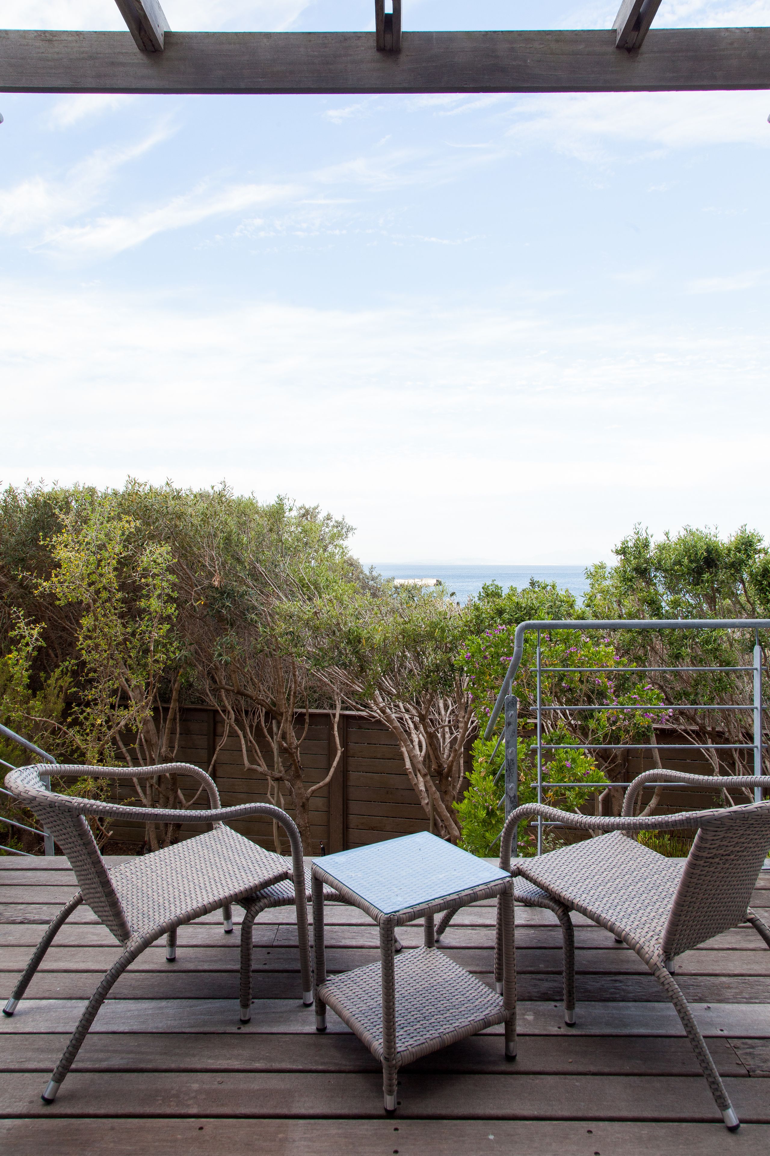 Uma área de estar coberta ao ar livre, com cadeiras de vime e uma mesa, com vista para um jardim e o mar.