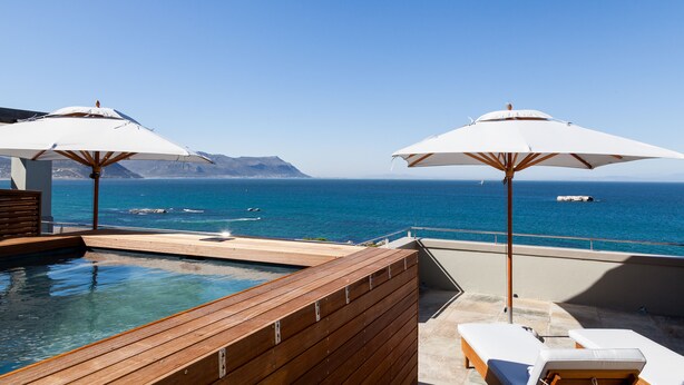 Zona de piscina en la azotea con cubierta de madera, sillones de descanso y sombrillas, con vistas a un mar azul cristalino y montañas a lo lejos.