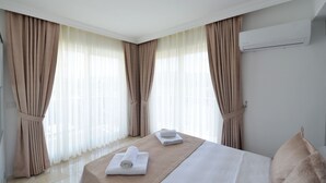 Habitación de hotel con una cama bien hecha, dos toallas enrolladas, cortinas color beige y un equipo de aire acondicionado en la pared.