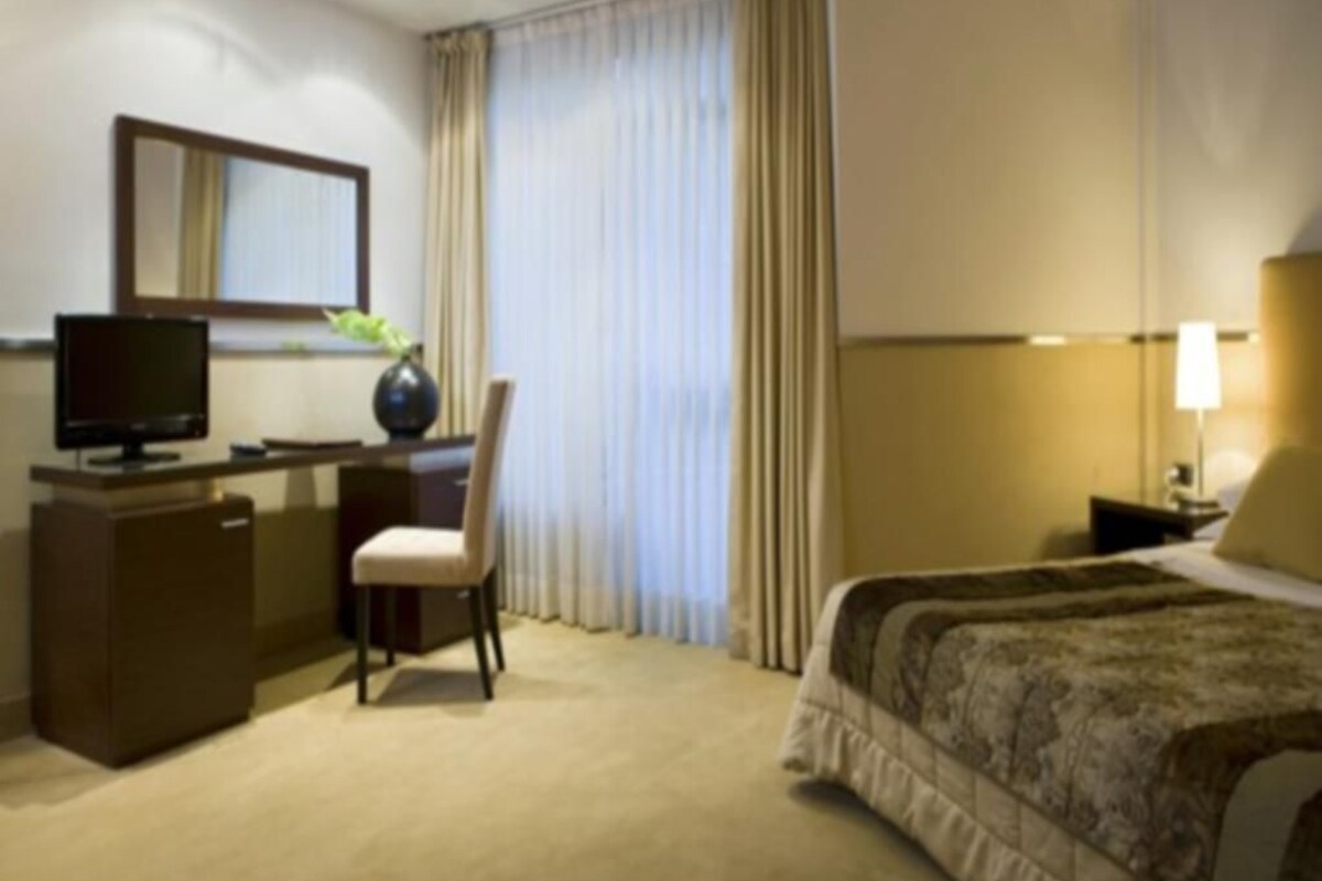 A hotel room with a bed, a desk with a television, a chair, and a window with curtains.