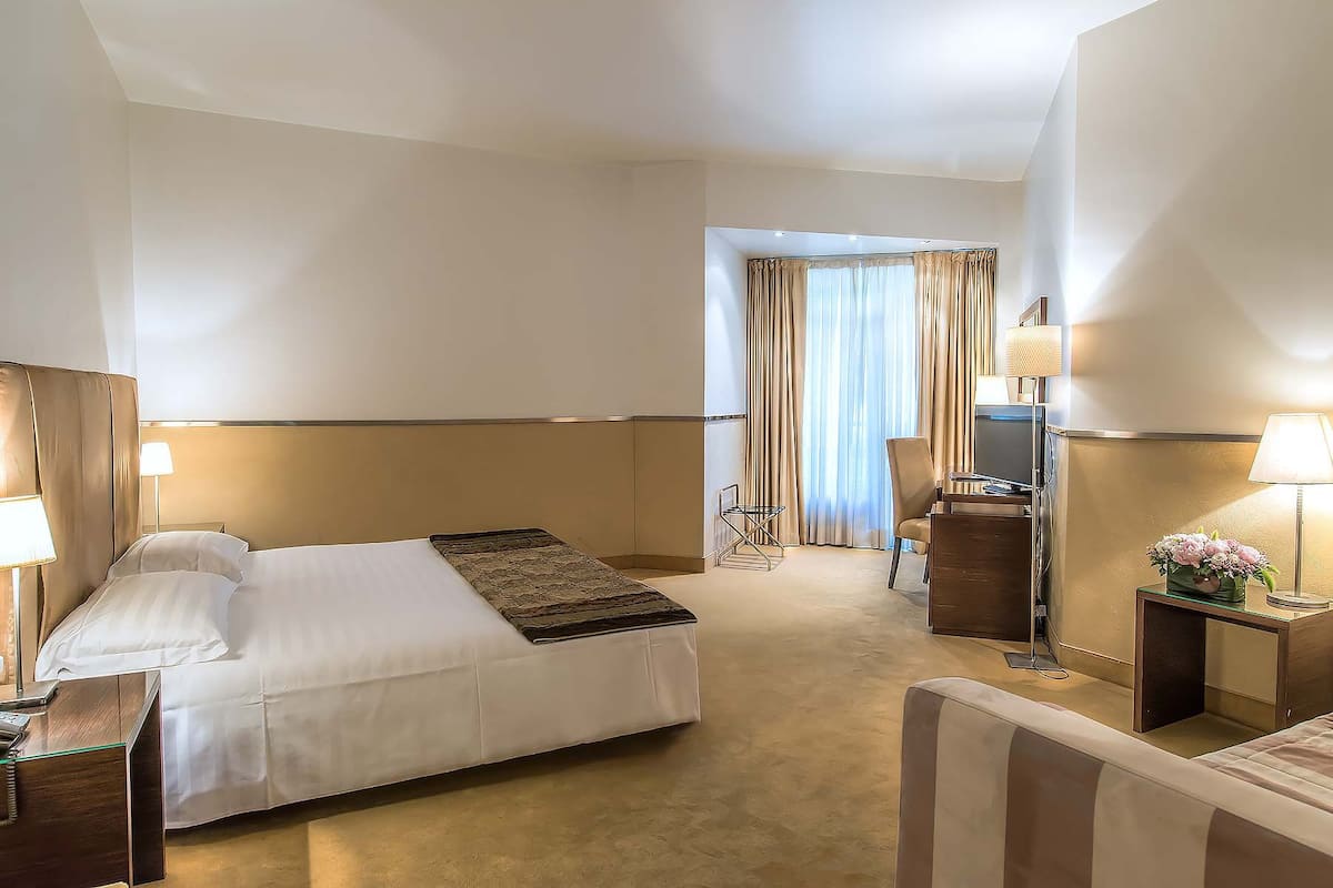 A hotel room with a bed, bedside tables, a desk, a chair, a TV, and a window with curtains.