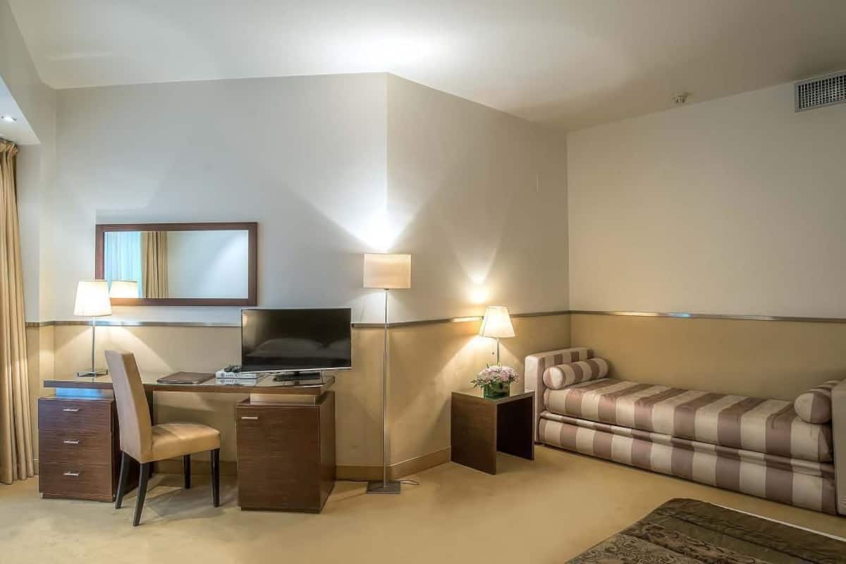 A hotel room with a desk, chair, flat-screen TV, two bedside lamps, a sofa, and a small side table with a flower vase.