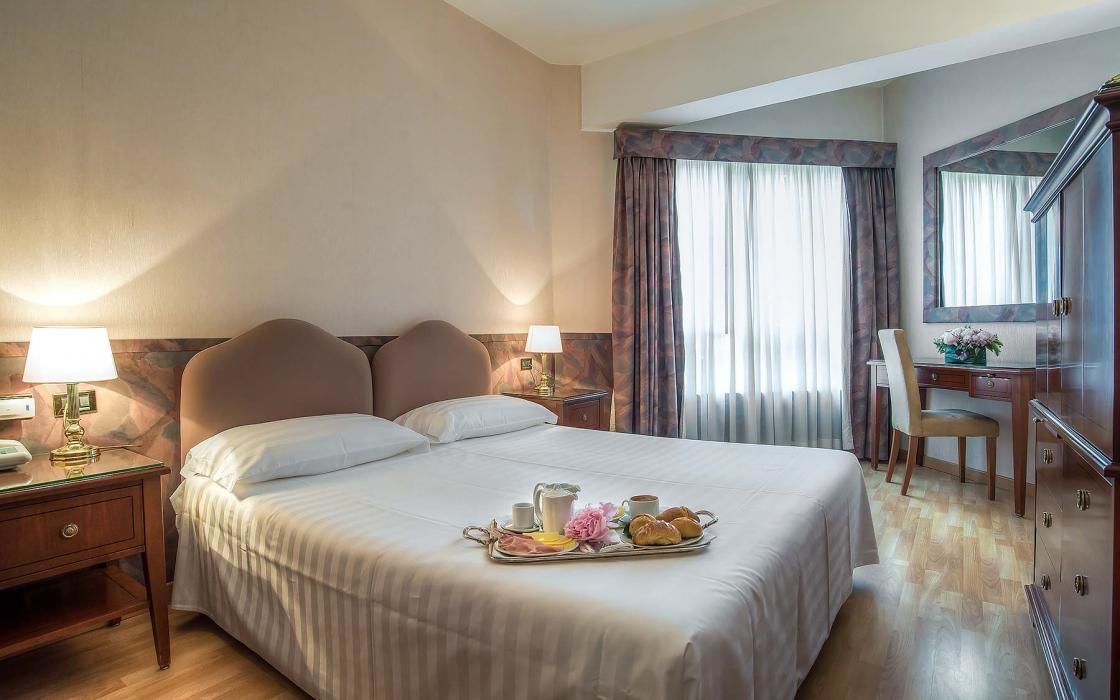 Camera d'albergo con un letto grande, lampade sui comodini, una scrivania con una sedia, una cassettiera e una finestra con tende.