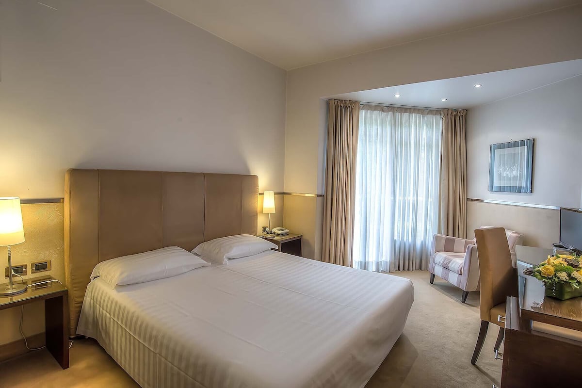 A hotel room with a large bed, two bedside tables with lamps, a desk with a chair, and a window with curtains.
