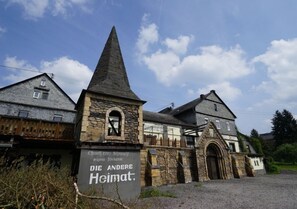 A historic building with a prominent stone tower and a sign reading "Die Andere Heimat."