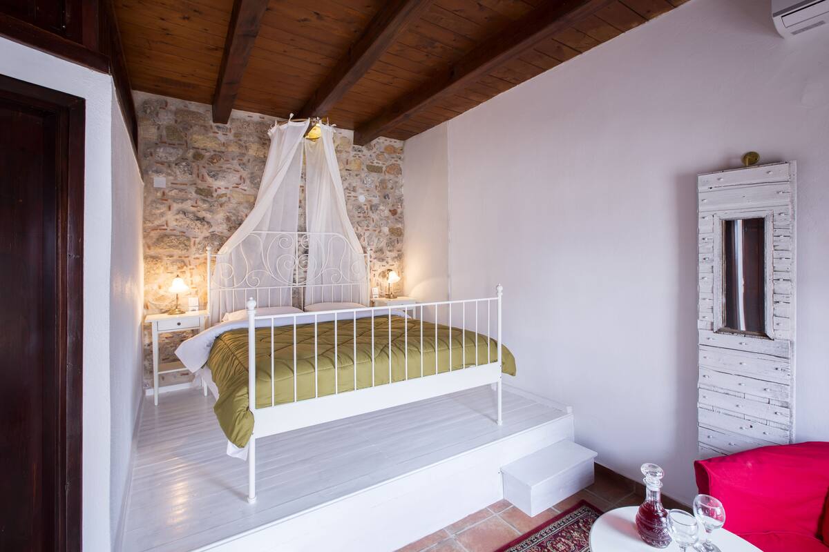 A bedroom with a white bed, a stone wall, a wooden ceiling, a nightstand, a lamp, a red chair, and a window with shutters.