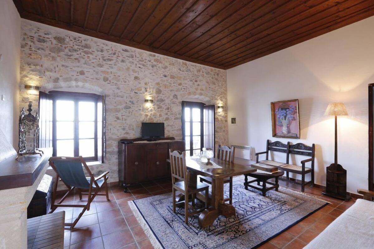 A rustic room with a stone wall, wooden ceiling, a dining table with chairs, a fireplace, and a lamp.