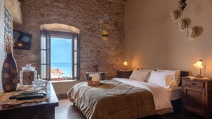 Un dormitorio con pared de piedra, una cama con manta color beige, una mesita de noche con lámpara y vista al mar a través de una ventana.