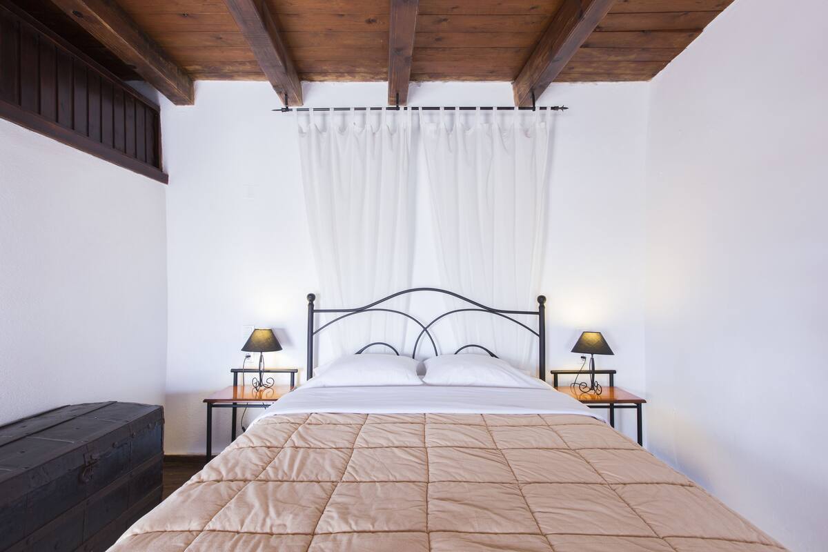 A bedroom with a bed, two bedside lamps, a wooden headboard, a wooden bench, and a white curtain.