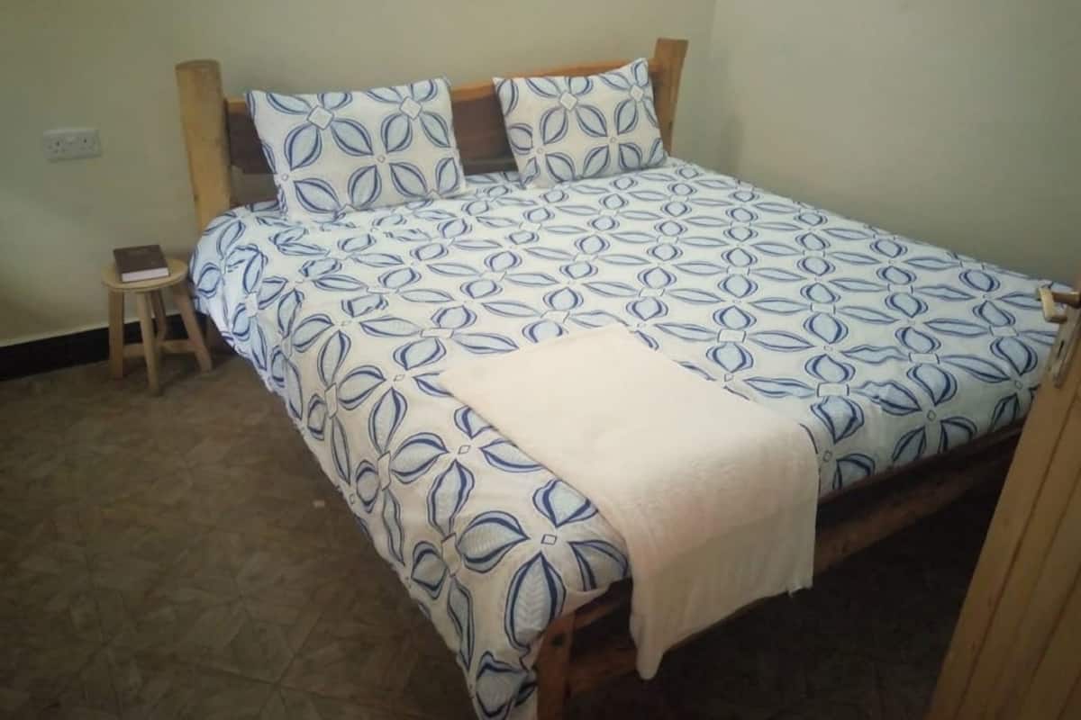 A bed with a patterned bedspread and pillows, a small wooden stool with a book, and a tiled floor.