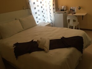 A neatly made bed with white linens, a small desk with a kettle and cups, and a chair.
