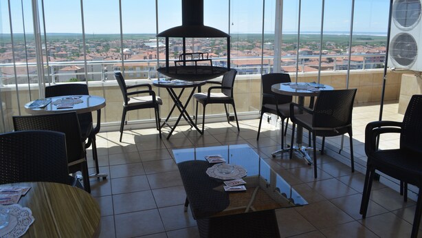 Terraza soleada con vistas a un paisaje urbano, amueblada con mesas y sillas, y una parrilla en el centro.