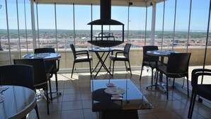 Un espacio comedor al aire libre iluminado por el sol, con varias mesas y sillas, una parrilla negra en el centro y una vista panorámica de un paisaje urbano.