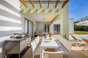 Un espacio moderno para comer al aire libre con una parrilla, asientos y vistas a una piscina.