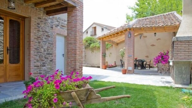 A courtyard with a stone wall, a wooden door, a covered patio area with a table and chairs, and a wheelbarrow with flowers.